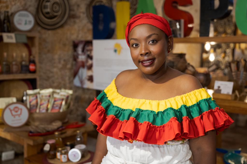 Mujer afrocolombiana con vestido colorido de hombros descubiertos con volantes amarillos, verdes y rojos, frente a una mesa con frascos, bolsas de productos y platos decorativos. Al fondo se observan letras grandes que forman la palabra "DASP" y un panel de texto desenfocado.