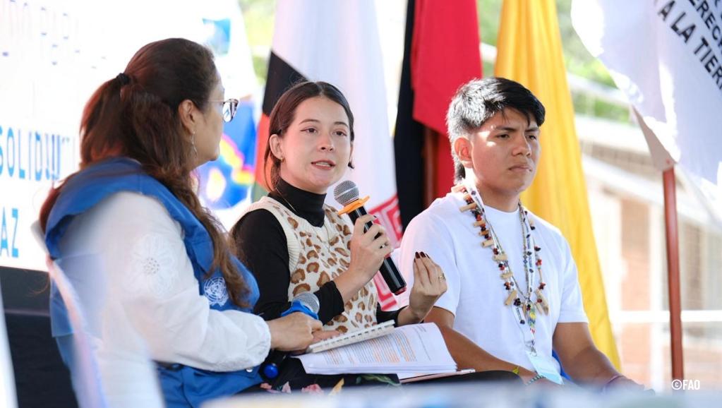 Jóvenes líderes indígenas y representantes de organizaciones internacionales dialogan sobre derechos territoriales y paz en el X Foro Global de la Tierra, con banderas y mensajes de solidaridad de fondo.