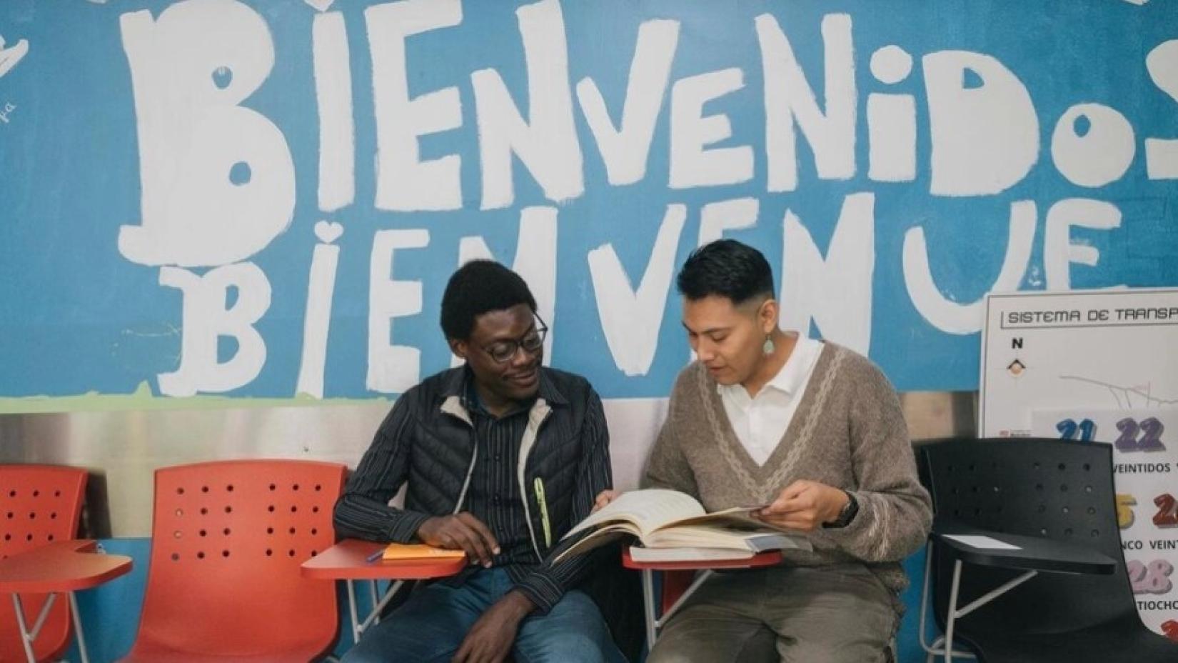 Dos hombres jóvenes conversan mientras revisan un libro en un espacio educativo. Al fondo, una pared azul con el mensaje “Bienvenidos” y “Bienvenue” escrito en grande, transmitiendo un ambiente de acogida e inclusión. Imagen relacionada con la labor de integración y acompañamiento de Naciones Unidas en Colombia. © ACNUR/Jeoffrey Guillemard