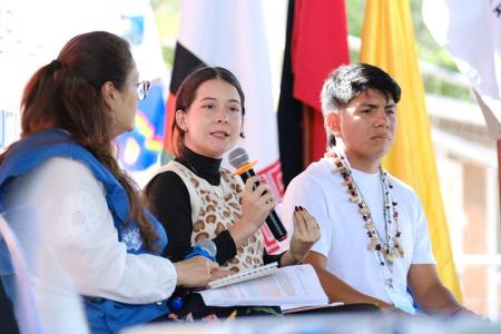 Jóvenes líderes indígenas y representantes de organizaciones internacionales dialogan sobre derechos territoriales y paz en el X Foro Global de la Tierra, con banderas y mensajes de solidaridad de fondo.