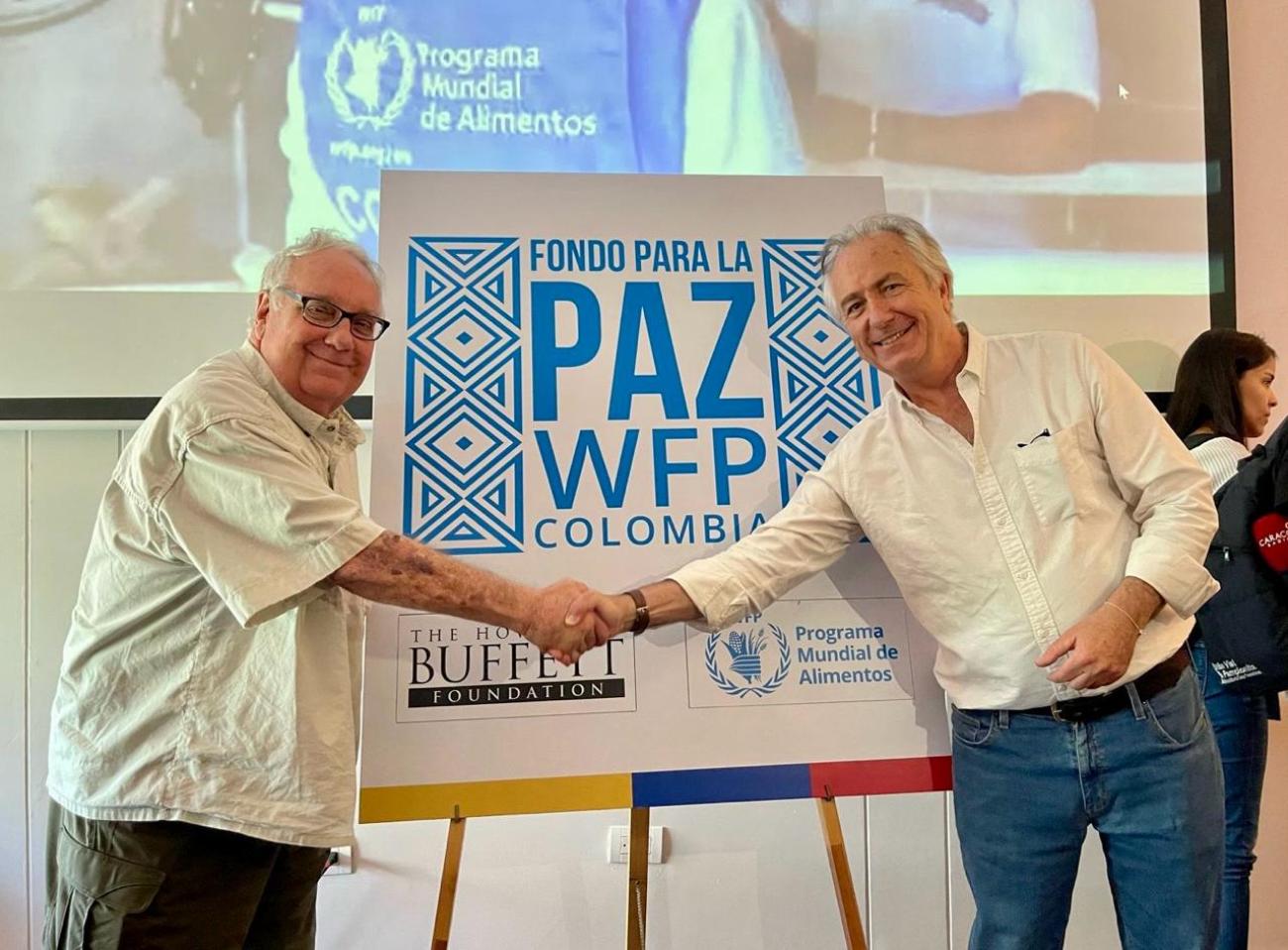 El filántropo estadounidense Howard Buffet (izquierda) y Carlo Scaramella, Director de País de WFP Colombia (derecha) estrechan la mano delante de un cartel que dice Fondo para la Paz WFP Colombia.