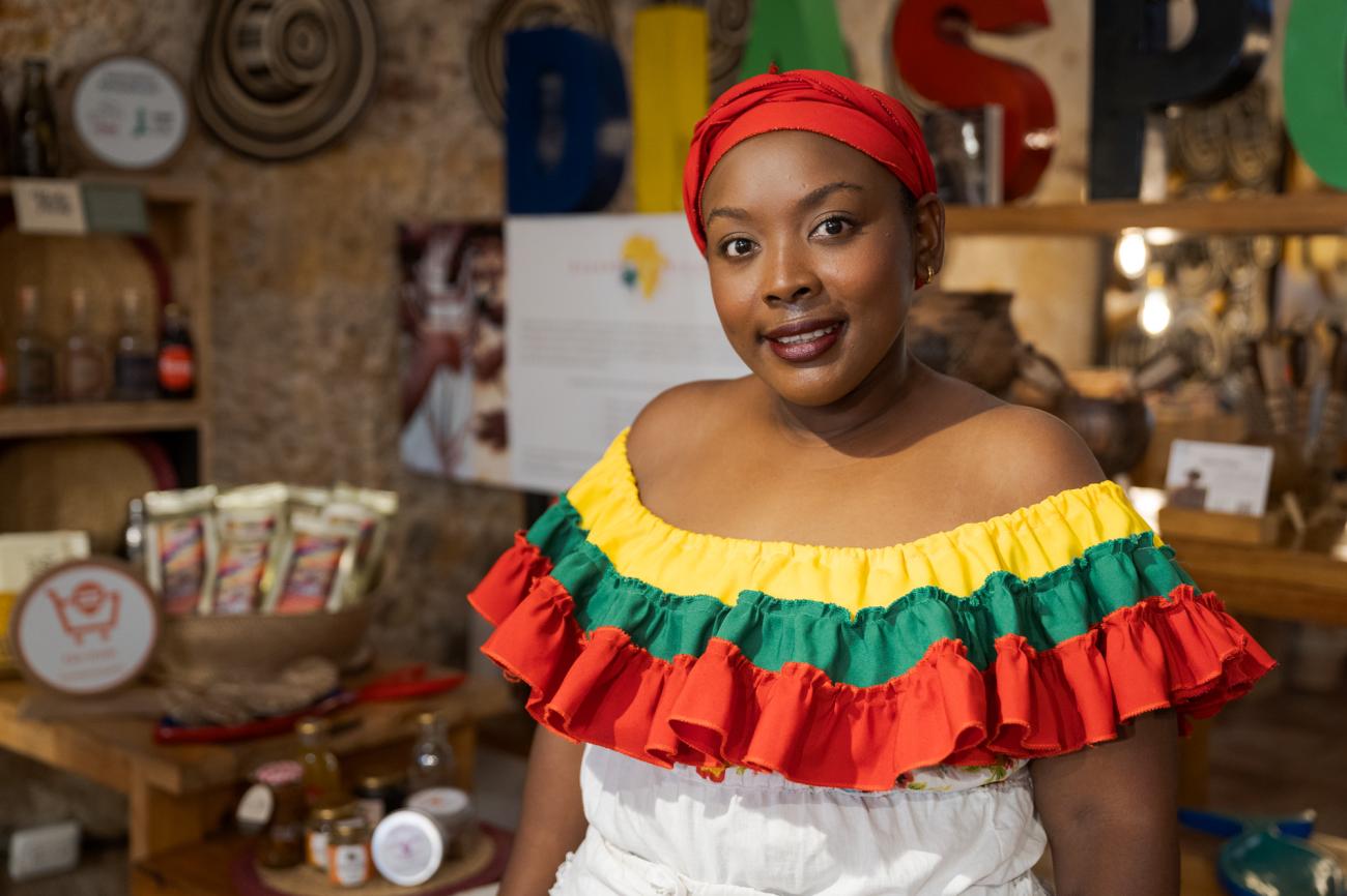 Mujer afrocolombiana con vestido colorido de hombros descubiertos con volantes amarillos, verdes y rojos, frente a una mesa con frascos, bolsas de productos y platos decorativos. Al fondo se observan letras grandes que forman la palabra "DASP" y un panel de texto desenfocado.