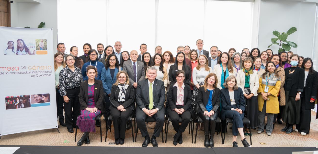 Foto grupal de unas 35 personas, hombres y mujeres, con vestimenta formal y de oficina, en una sala iluminada con paredes blancas. A la izquierda, un cartel con el texto “Mesa de género de la cooperación internacional en Colombia” y fotos de personas colaborando.