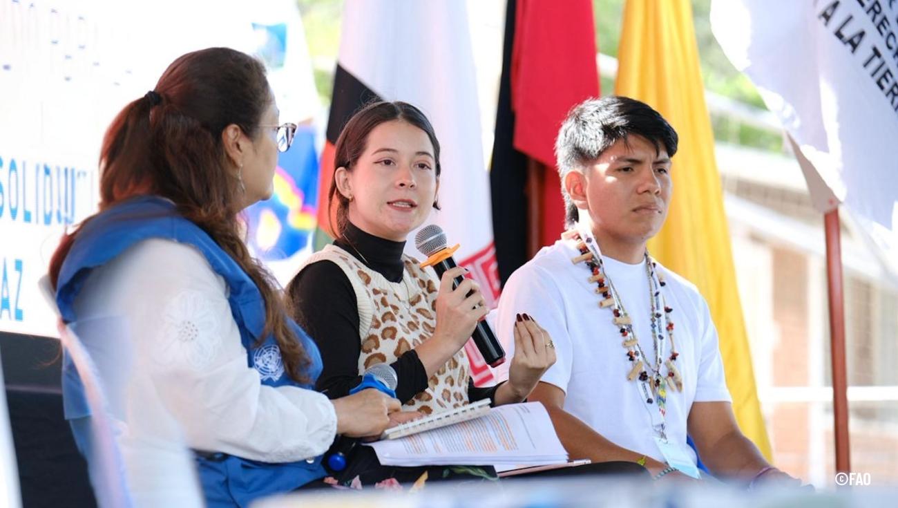 Jóvenes líderes indígenas y representantes de organizaciones internacionales dialogan sobre derechos territoriales y paz en el X Foro Global de la Tierra, con banderas y mensajes de solidaridad de fondo.