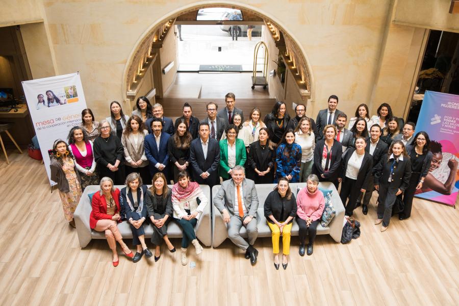 Fotografía grupal de aproximadamente 40 personas, hombres y mujeres, vestidas formalmente y reunidas en el vestíbulo de un edificio elegante con piso de madera. Algunas personas están sentadas en sillones al frente, mientras que el resto está de pie detrás. Se observan pancartas informativas a los lados.