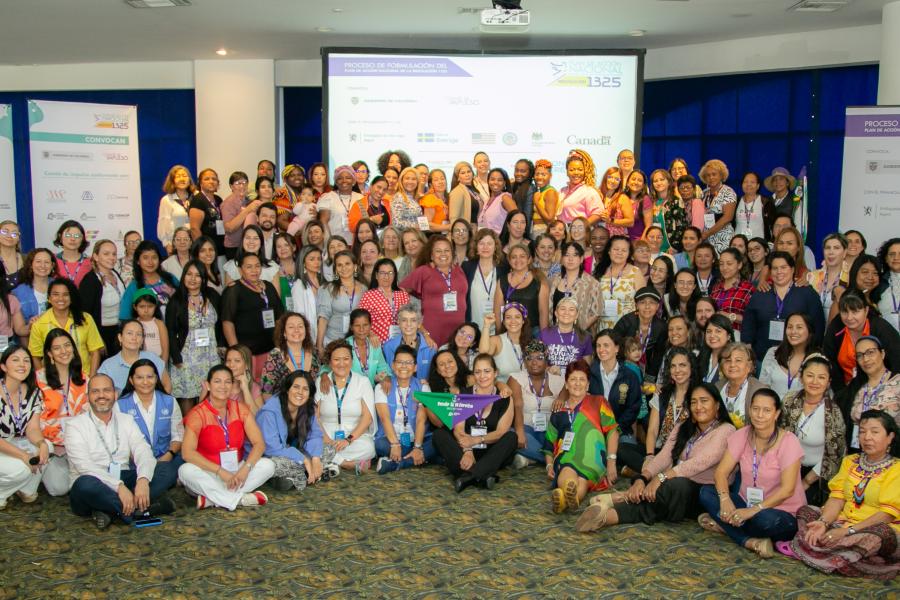 Fotografía grupal de más de 100 mujeres reunidas en un salón de conferencias, muchas de ellas sentadas en el suelo o en sillas, y otras de pie. Al fondo se ven pantallas grandes con logotipos y pancartas informativas, indicando un evento formal o conferencia. Todas sonríen y posan para la cámara.