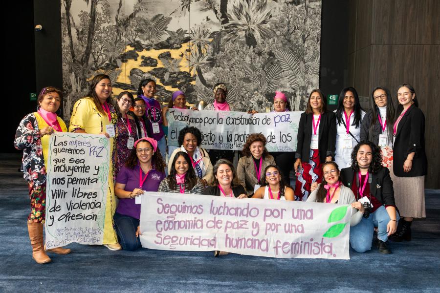 Grupo de aproximadamente 20 mujeres posando con pancartas en un espacio interior con una gran obra de arte en blanco y negro al fondo. Las pancartas tienen mensajes en favor de la paz, la seguridad y los derechos de las mujeres desde una perspectiva feminista. Algunas mujeres visten prendas moradas y fucsias, símbolos del movimiento feminista.