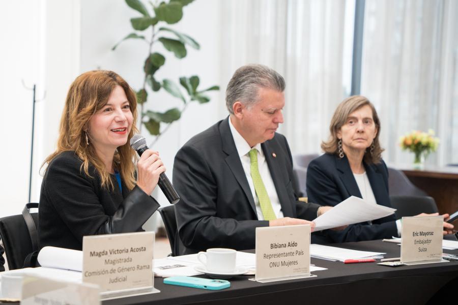 Tres personas sentadas en una mesa de conferencia con carteles de nombre: Magda Victoria Acosta (Magistrada Comisión de Género Rama Judicial), Bibiana Aido (Representante País ONU Mujeres) y Eric Mayoraz (Embajador Suizo). La persona a la izquierda habla por micrófono. Sobre la mesa hay papeles, tazas de café y una planta al fondo.