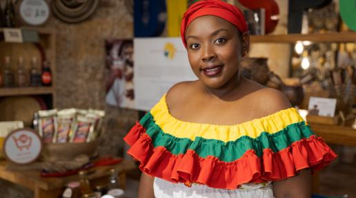 Mujer afrocolombiana con vestido colorido de hombros descubiertos con volantes amarillos, verdes y rojos, frente a una mesa con frascos, bolsas de productos y platos decorativos. Al fondo se observan letras grandes que forman la palabra "DASP" y un panel de texto desenfocado.