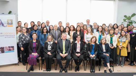 Foto grupal de unas 35 personas, hombres y mujeres, con vestimenta formal y de oficina, en una sala iluminada con paredes blancas. A la izquierda, un cartel con el texto “Mesa de género de la cooperación internacional en Colombia” y fotos de personas colaborando.